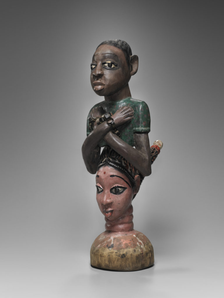 A wooden sculpture with the bust of a Black man with chains around his wrists and the bust of a South Asian woman below him.