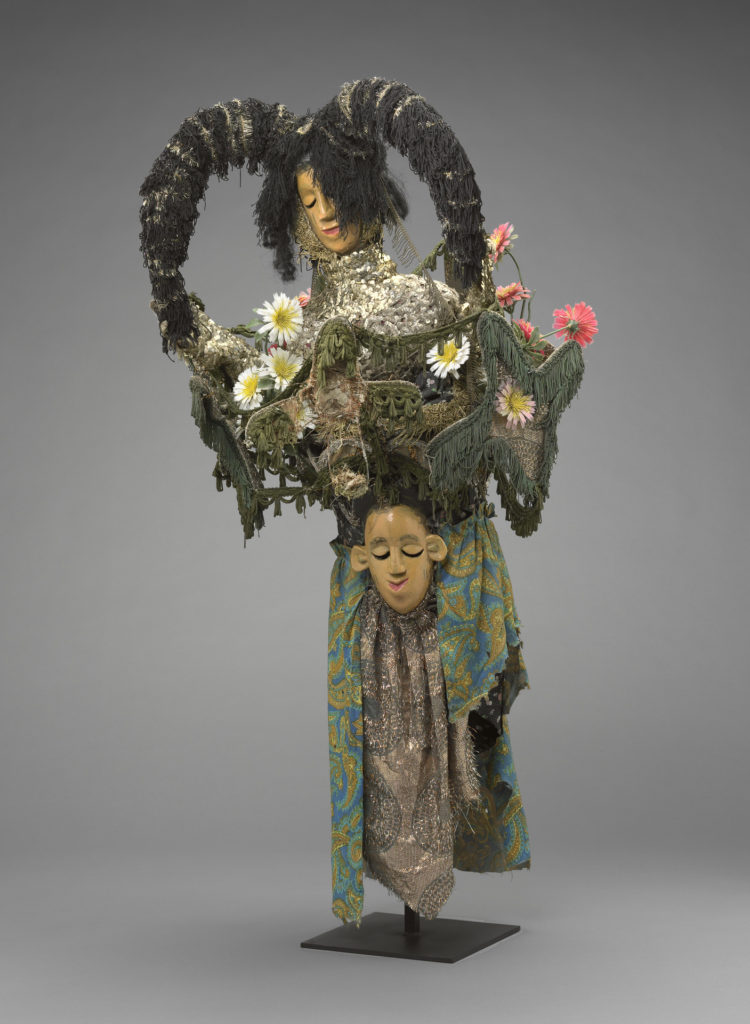 A masquerade costume with two faces, plastic flowers, multicolored textiles, and two large horns protruding off one of the faces.