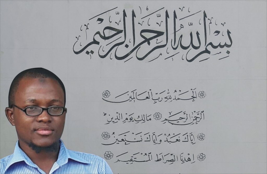 Ibrahim Jimoh is shown standing before his ijazah that displays Surat Al-Fatihah. The first verse (basmalah) is written in the Thuluth script while the remaining verses are written in a smaller Naskh script.