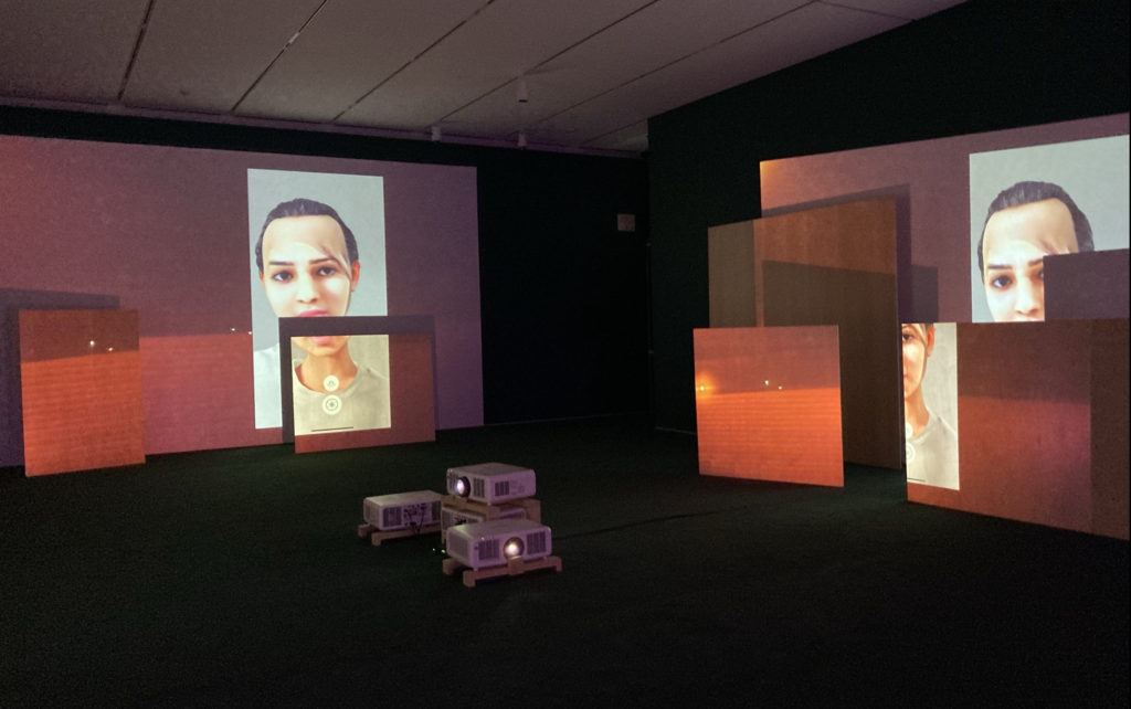 Image of a dark gallery room with a green carpet. 4 projectors are set upon wooden frames in the middle of the image, on the floor. On one wall is a projected image of walls dividing Palestinians from each other; it is projected onto wooden slabs overlapping each other. Additionally, a digitally altered image of a young Palestinian is against this backdrop. The same is projected onto the other wall, with a digital image of the scarred Palestinian child –with no gendered features.