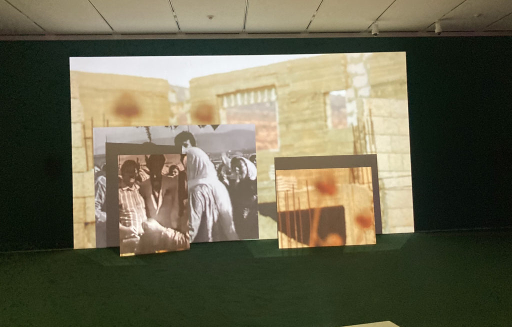 Image of a dark gallery room with a green carpet. Still of video being projected onto boards. Note that the wooden boards the images are projected onto are overlapping and are of different sizes. In the foreground, black and white footage is projected of Palestinians; the footage is historical and is of a crowd. The background is in color and features a demolished Palestinian house, with nothing but the walls remaining. Exterior of the building is masoned stone and the interior is grey concrete and thick construction wires.