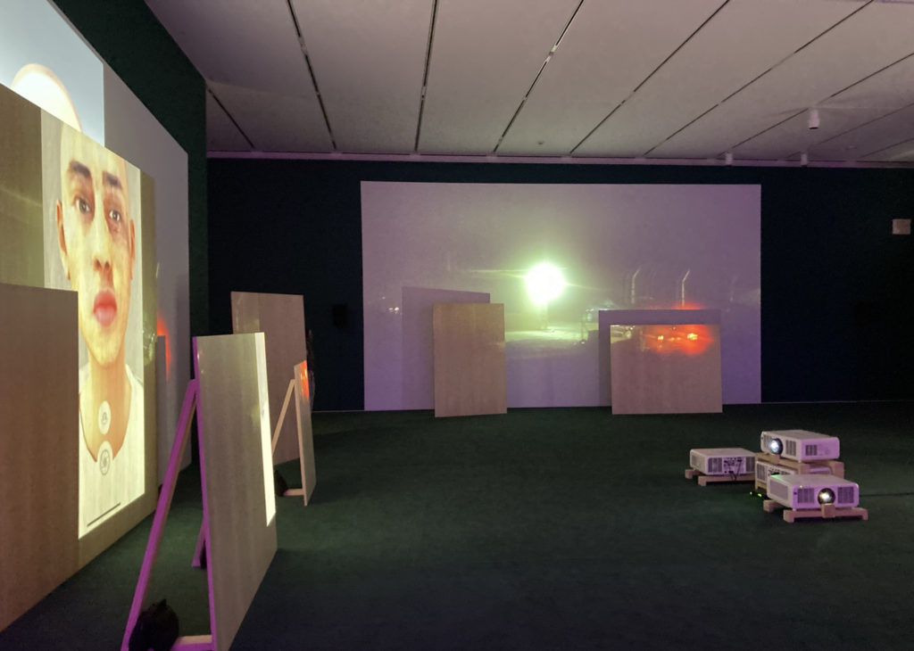 Image of a dark gallery room with a green carpet. 4 projectors are set upon wooden frames in the middle of the image, on the floor. On one wall is a scarred Palestinian child with no gendered features; the image is digitially altered. The other is of a wire fence at night with security vehicles in front of it.