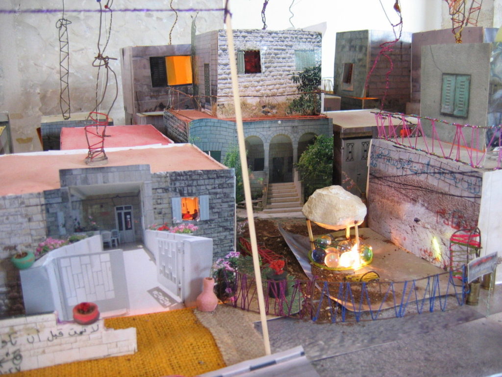Image of a landscape, representing Qalandia, of miniature buildings made of grey brick and cement with doors, windows and wires sticking from the tops. 