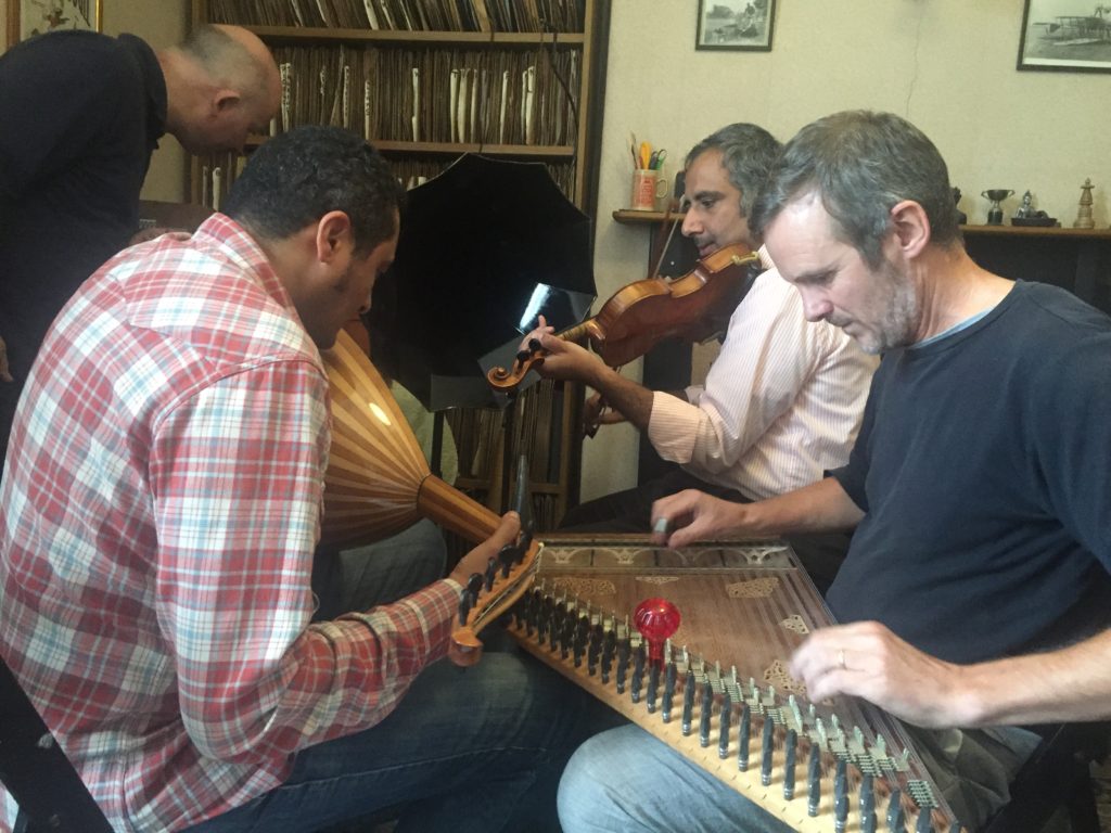 Photo of Martin Stokes, Tarik Beshir and Ahmad al-Salhi seated in front of a large phonograph horn playing qanūn, ‘ūd and violin respectively. Duncan Miller, in the background, is leaning over phonograph attentively.
