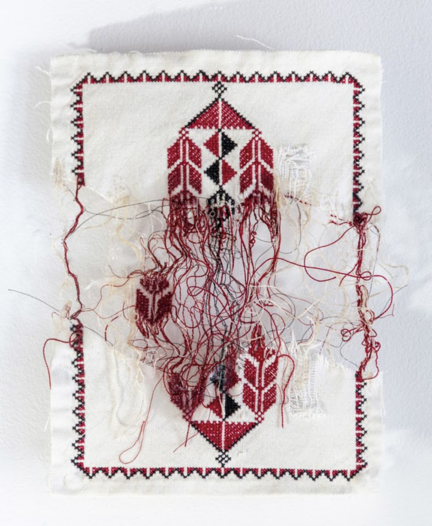 A piece of Palestinian embroidery in black and red: the embroidery piece is damages in the middle revealing a messy of threads. The piece has a border and is set on a white background.