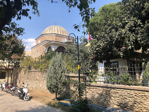 A view of the library grounds from a diagonal angle shows the library, including its garden with fruit and magnolia trees, bordered by a brick wall and iron grates. On the other side of the brick wall is the grounds of the neighboring Ibn Melek shrine where a lamp post, a few parked motorbikes and a small cypress tree are visible in the summer heat.
