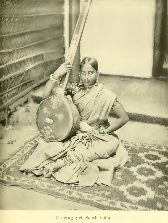  A black and white image of a young woman sitting holding either a sitar or veena on her lap. Her right hand is tuning the instrument while her other hand is on her waist. She is sitting on a decorative carpet and is herself adorned with jewels like bangles, nose rings, earrings, and necklaces. Her sari is neatly tucked out enough to showcase her anklets. She has neatly parted hair.
