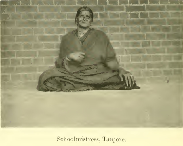 Alternative text: A grey image of an elderly woman sitting draped in a sari with one hand holding the sari and another resting on her left knee. She is resting against the brick wall. She is smiling. She is wearing a bindi. Her outfit is very simple as it is possibly a school uniform. She is not wearing any jewellery except one bangle.
