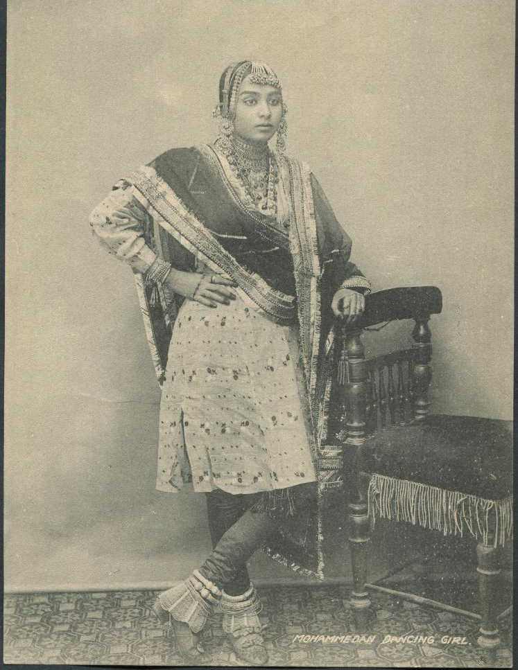 Black and white of an unnamed dancing girl who is identified as being Muslim. She is standing cross legged with one hand resting on a decorative plush chair next to her and the other hand on her waist. She is wearing a long kurta (shirt) with a V-neck collar. Her pants are tight with netted frills at the bottom giving an almost bell-bottom type of look. She is wearing huge (possibly silver) anklets and her shoes have a slight heel attached. She has henna on her hands. She is wearing immaculate and elaborate jewellery including, rings, bangles, earrings, multiple lengths of necklaces, hair jewellery, and a maang tikka (headpiece that goes in the middle of your forehead).
