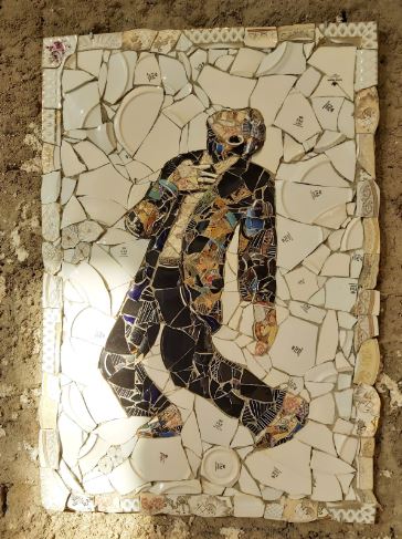 A rectangular collage made of porcelain pieces that together convey an image of a man in a black suit with his head tilted upwards, his right hand on his chest and his legs folded backwards. The rest of the collage is a white background with some of the white pieces displaying the Fathi Mahmoud logo. The background of the artwork is a beige cement wall.