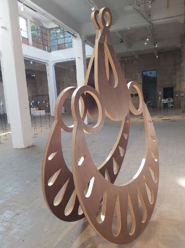 A plywood jewelry pendant sculpture is shown at an angle and shaped like a crescent, lying on its side, from the bottom with holes shaped like teardrops in it and two circles with holes in them are attached to each crescent tip. Between both circles, there is a teardrop-shaped structure with a teardrop-shaped hole in it and, at the top, there is another circle with a hole in it. The back of the pendant displays the same structure stuck to the front. The background shows a section of the room with white columns holding the windows above on the left. The ceiling is white. The beige-brown wall with a dark opening is shown on the right. The floor is beige-brown.